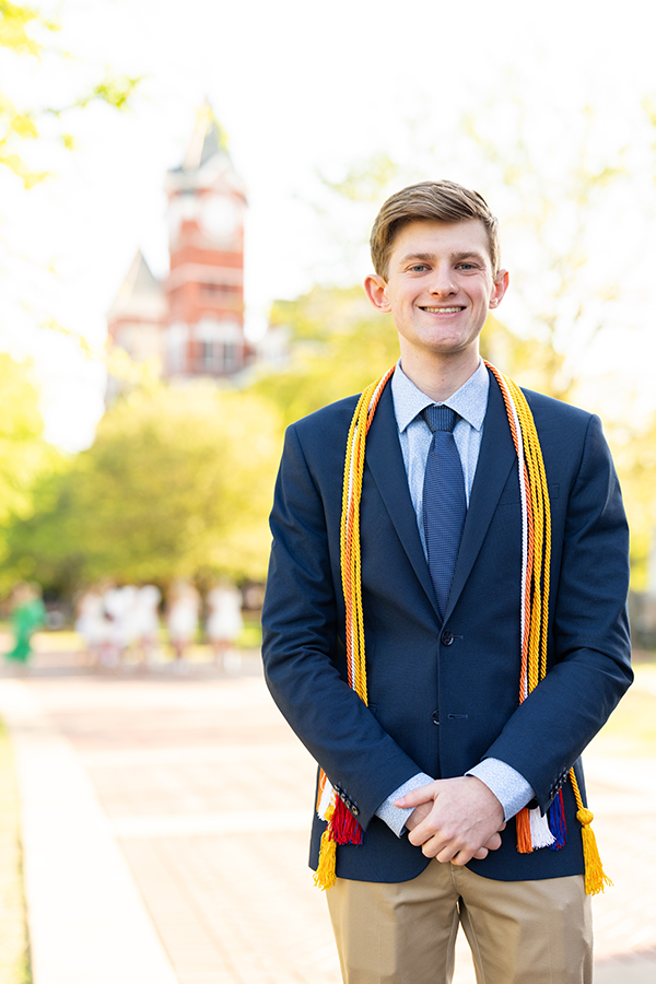 parker is standing outside with samford tower in the background. parker is wearing a blue suit with honor cords on