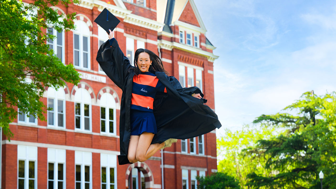 Hailey Tschetter jumping in a cap and gown in front of Samford Hall.